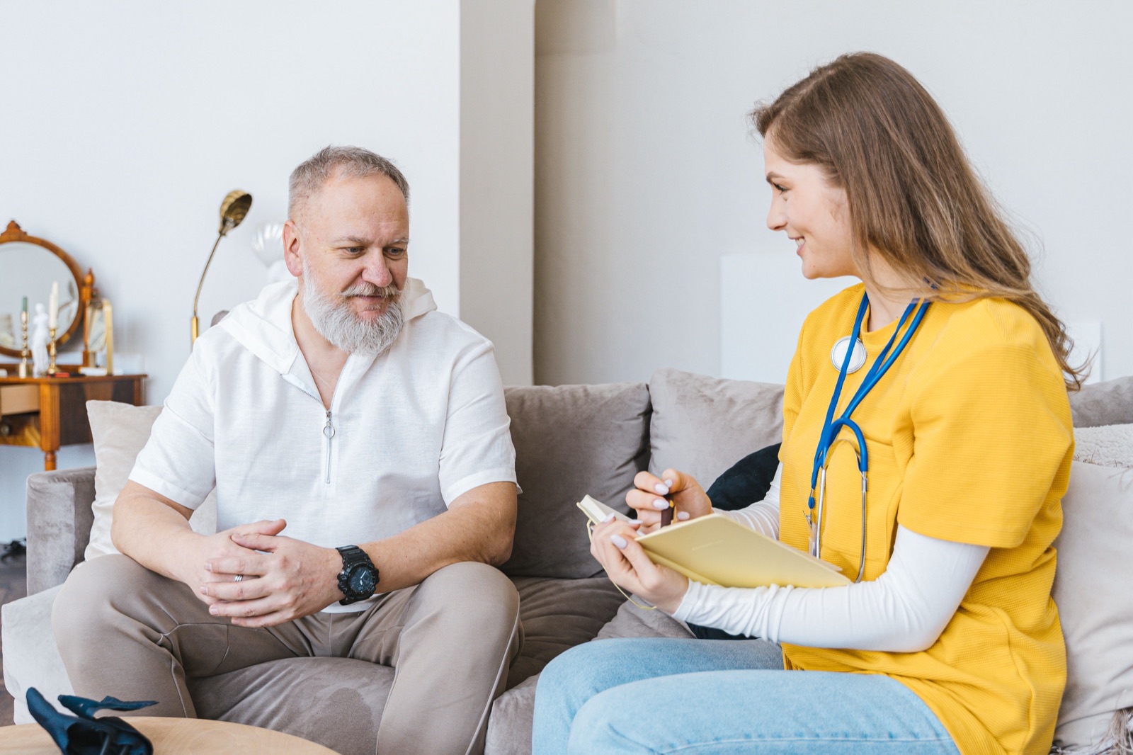A health worker with a stethoscope and notebook talks with an older bearded man on a sofa at home.