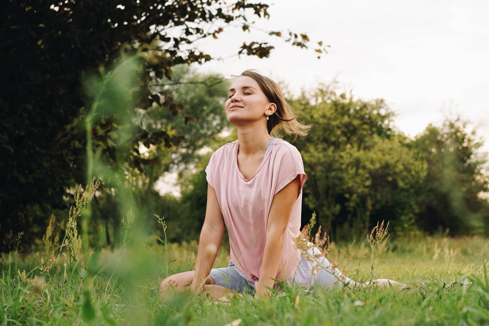 A woman in a pink T-shirt sits in tall green grass in a park, eyes closed and face lifted, breathing in the fresh air.