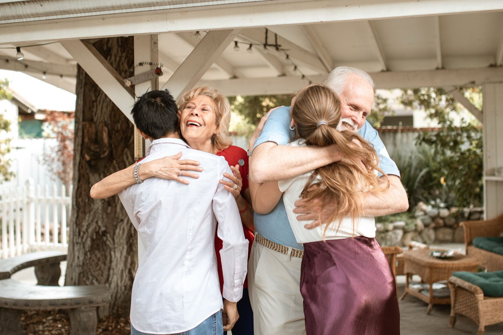 Under a shady porch roof, an older couple warmly hug two younger adults, the woman laughing with her eyes closed.