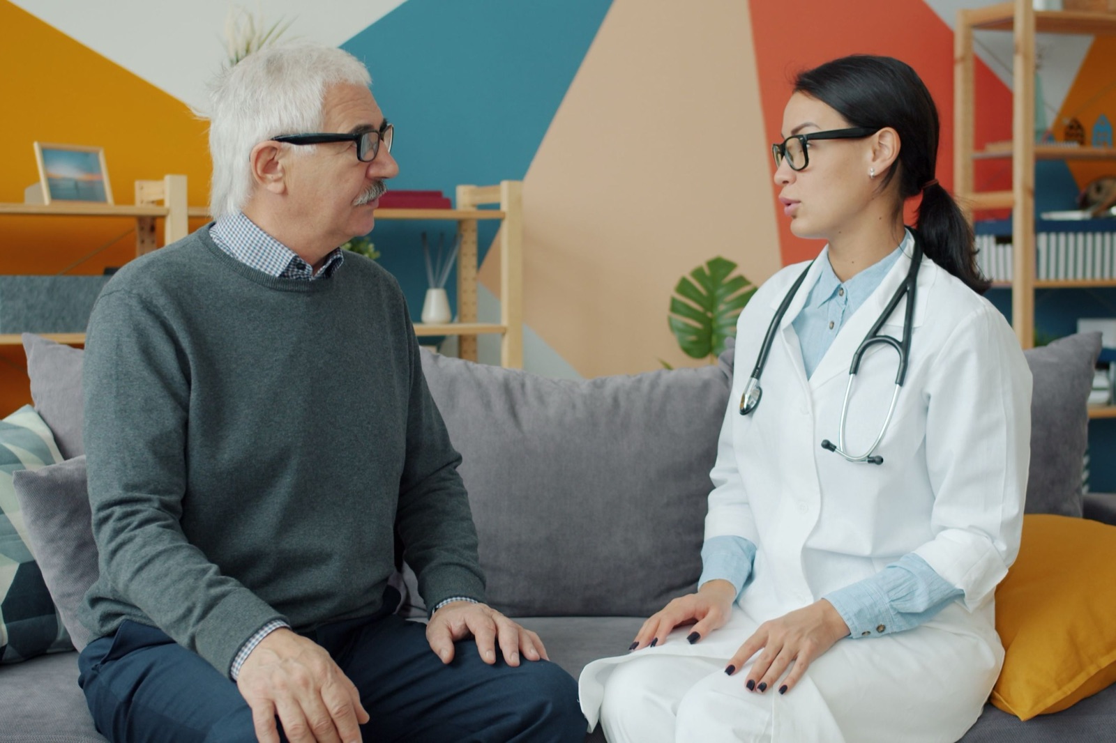 A doctor in a white coat and stethoscope talks face to face with an older man in glasses on a sofa in a bright, colorful office.