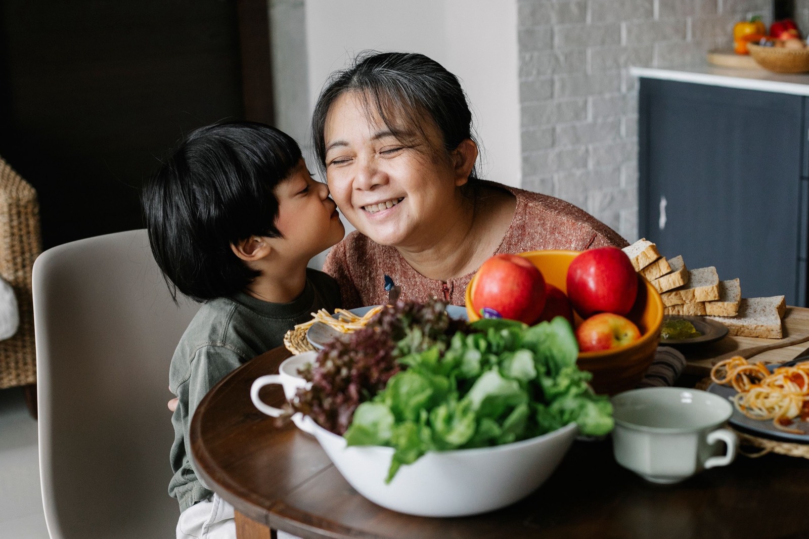 A young boy kisses his smiling grandmother on the cheek at a table set with a big bowl of salad greens, apples and bread.