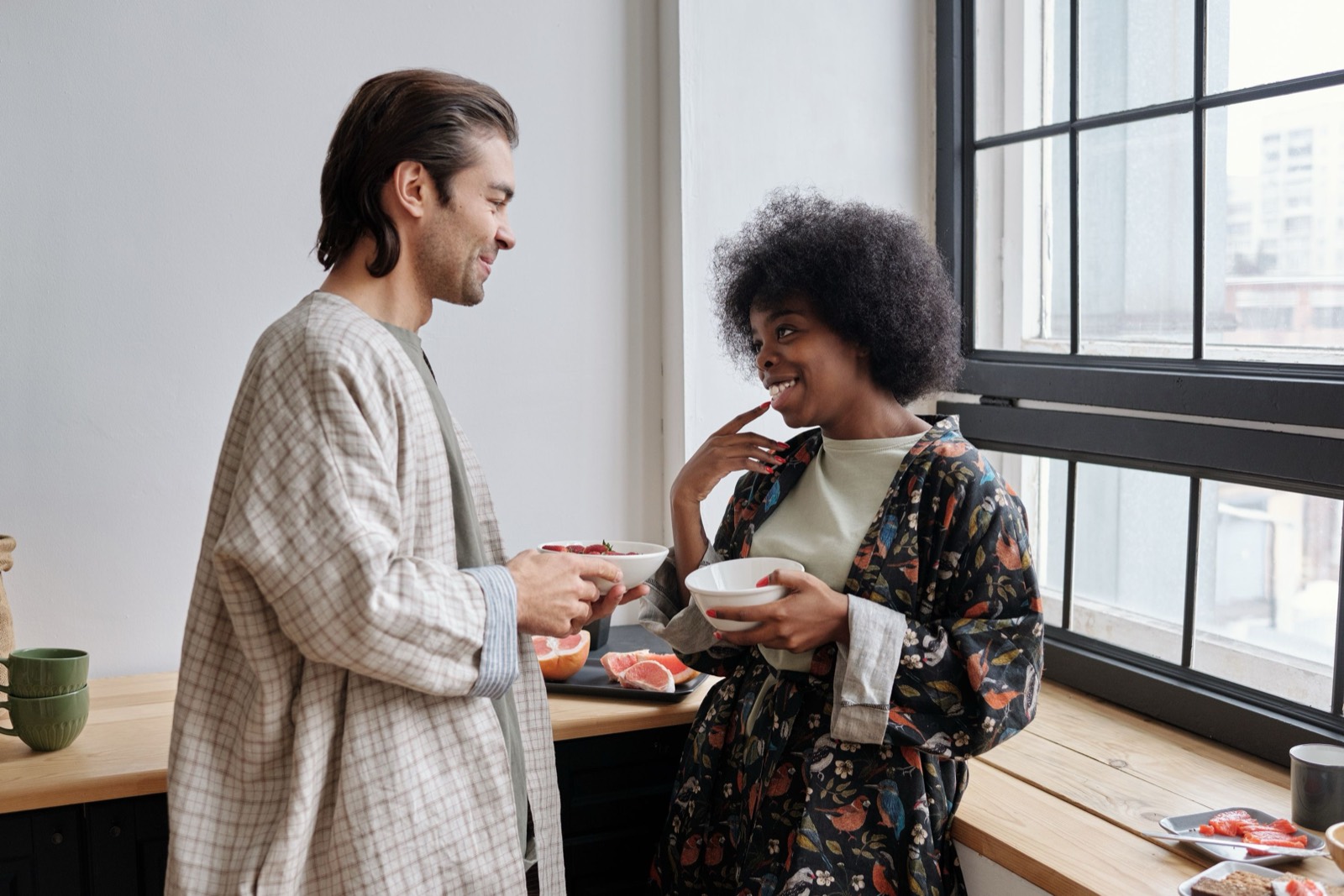 A couple chat and laugh by a sunny kitchen window, each holding a bowl of fruit, with sliced grapefruit on the counter.