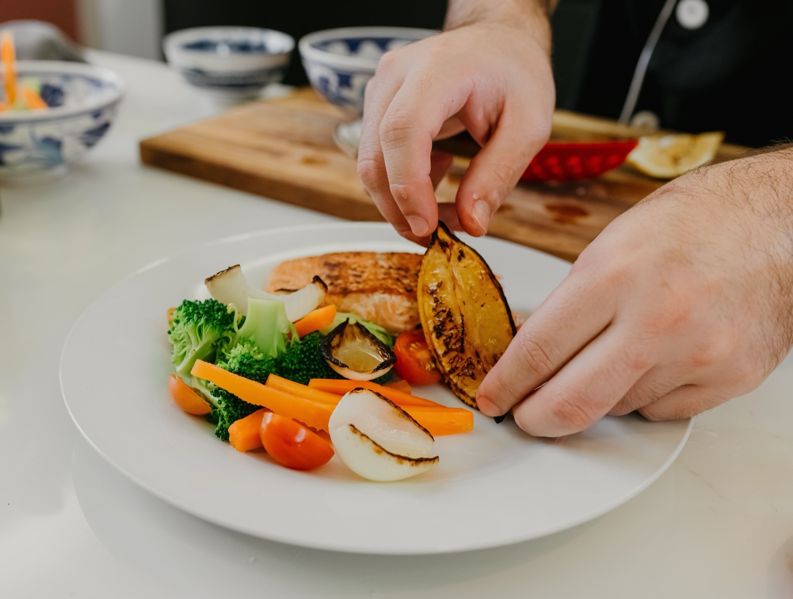 Hands place a grilled lemon half on a plate of salmon with broccoli, carrots, cherry tomatoes and roasted onion.