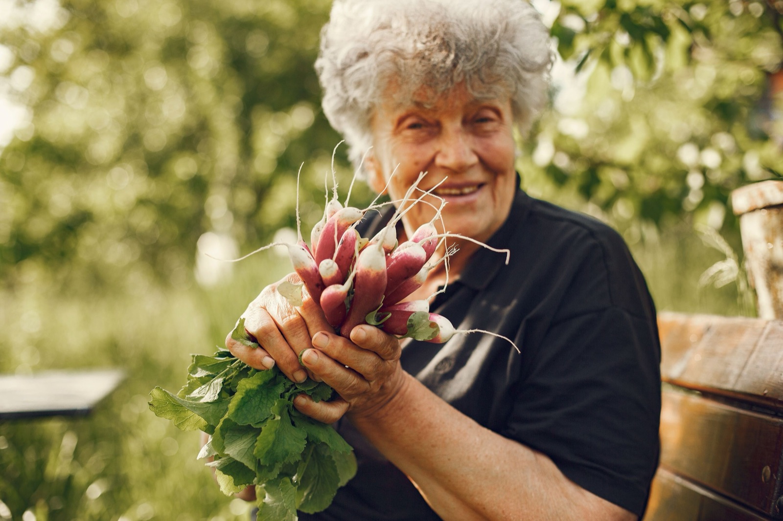 A smiling older woman with curly grey hair holds a big bunch of freshly pulled radishes in a sunny garden.