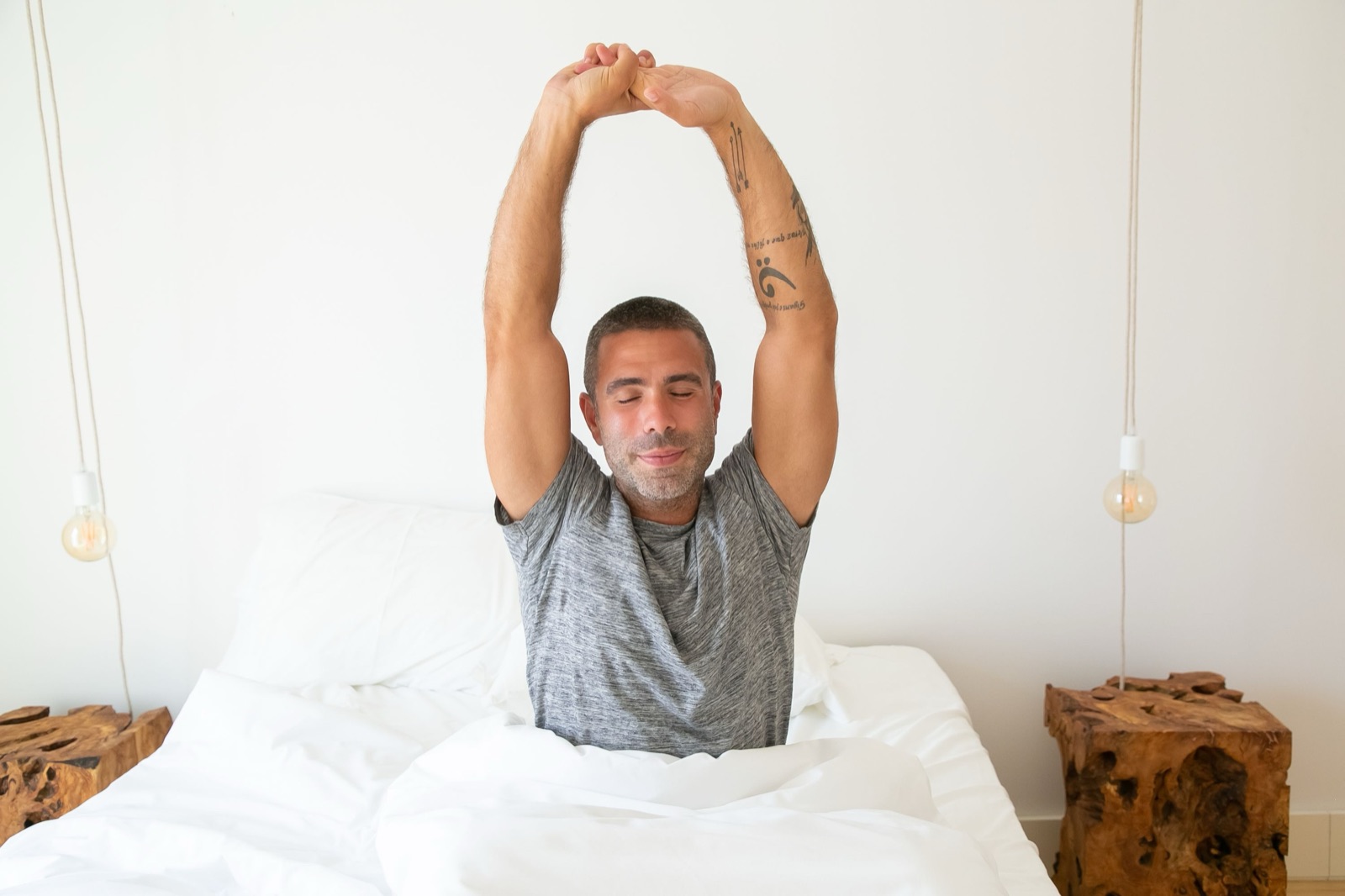 A man in a grey T-shirt sits up in a white bed and stretches both arms overhead, eyes closed and smiling.