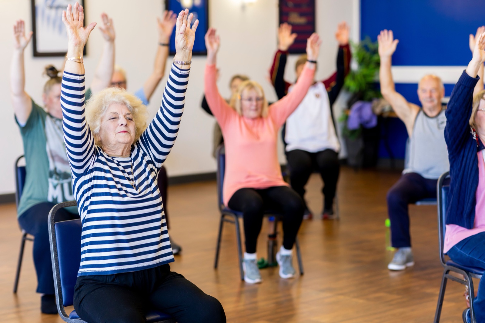 Older adults sitting on chairs in a bright hall raise both arms overhead during a group exercise class.