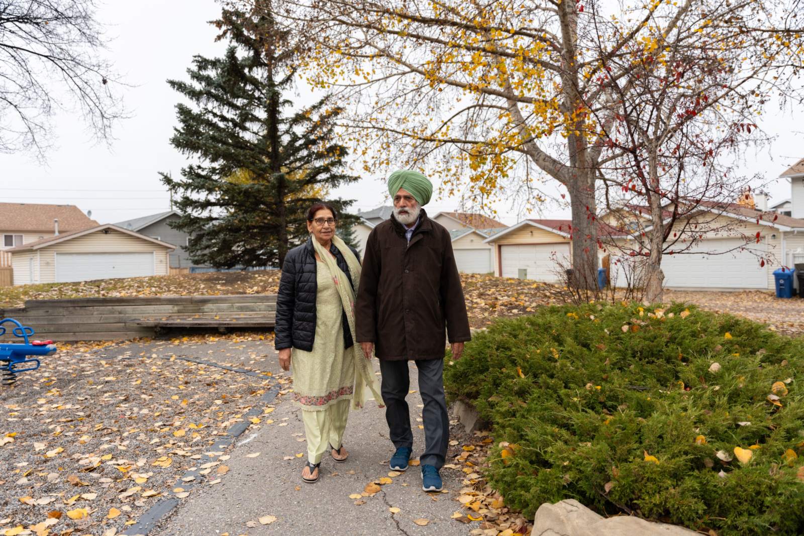 An older couple, the man in a green turban, walk side by side along a path covered in autumn leaves.